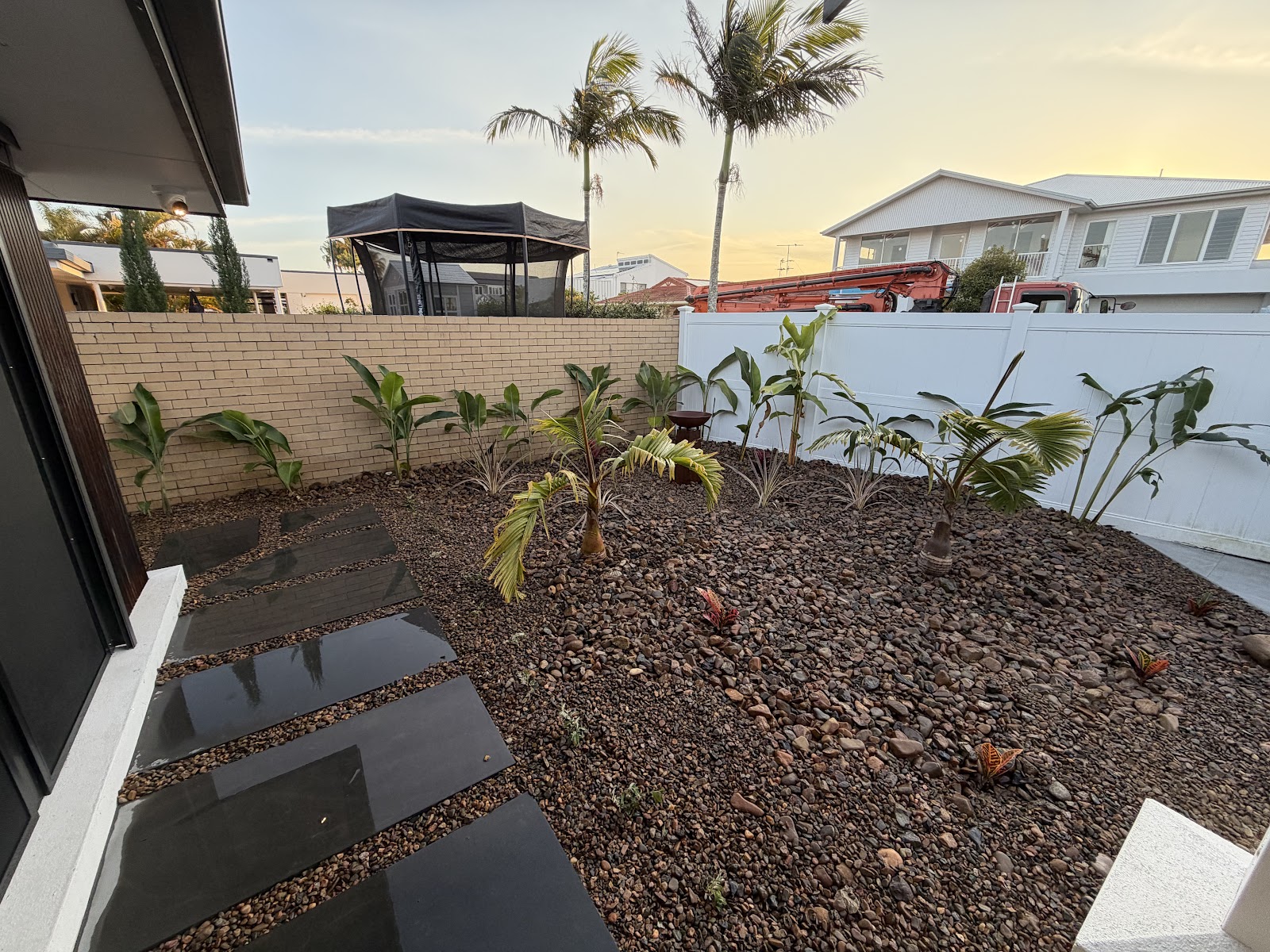 New pebble garden bed with stepping pavers and tropical planting beside a white fence
