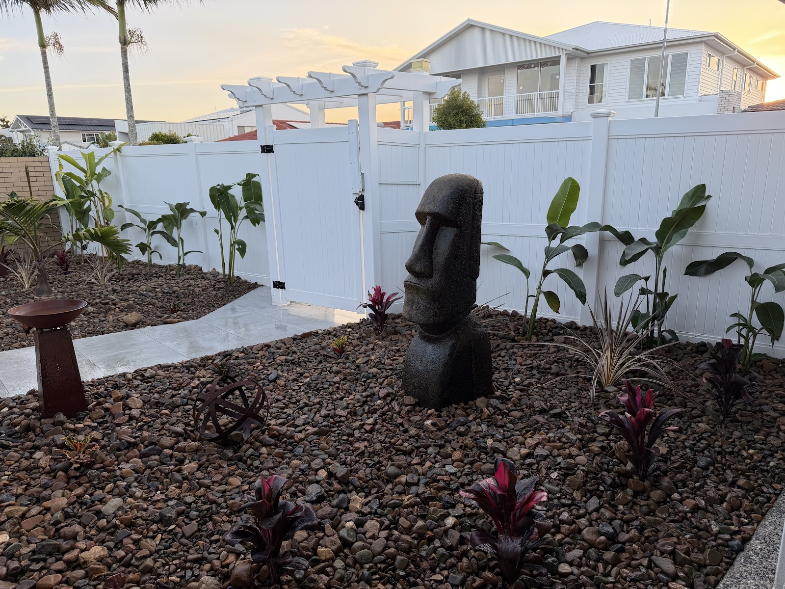 A finished tropical garden of river pebbles, cordylines and a stone head statue beside a white pergola gate